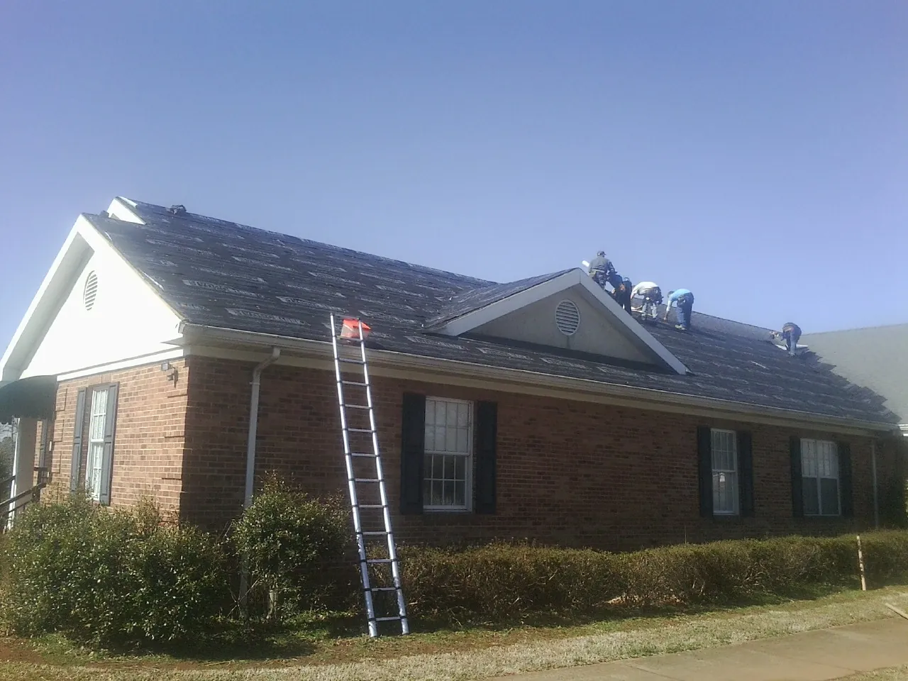 Crew tearing shingles off a brick ranch roof with a ladder set — Emergency Roof Repair work by our crew in Lowman